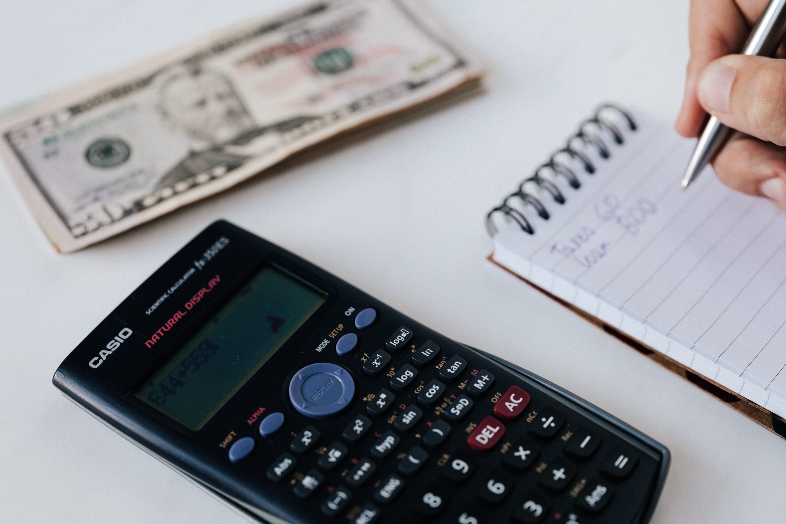 Casio calculator on a white table next to a stack of $50 bills while a person writes on a small notepad