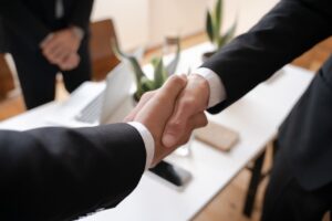 Two persons wearing black suit jackets are shaking hands while standing on a brown wooden floor near a white table in an office room