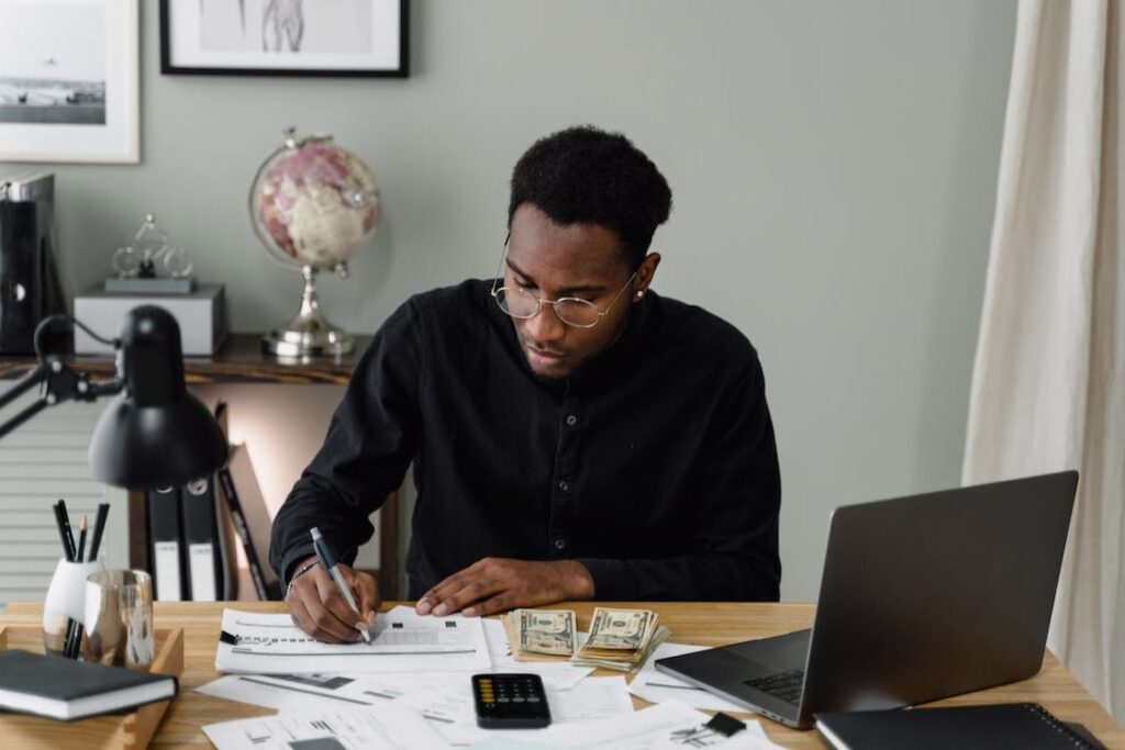 Man writing on a document with a calculator, paper bills and laptop on his table