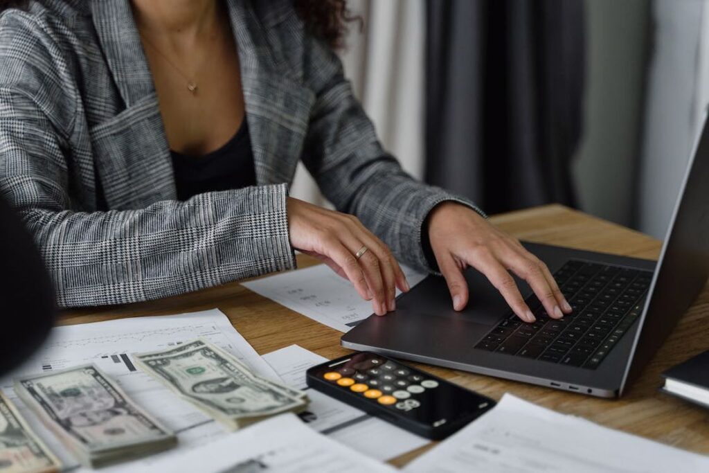 Woman wearing a gray suit jacket using her laptop while paper bills, papers and a calculator are laid out on her table