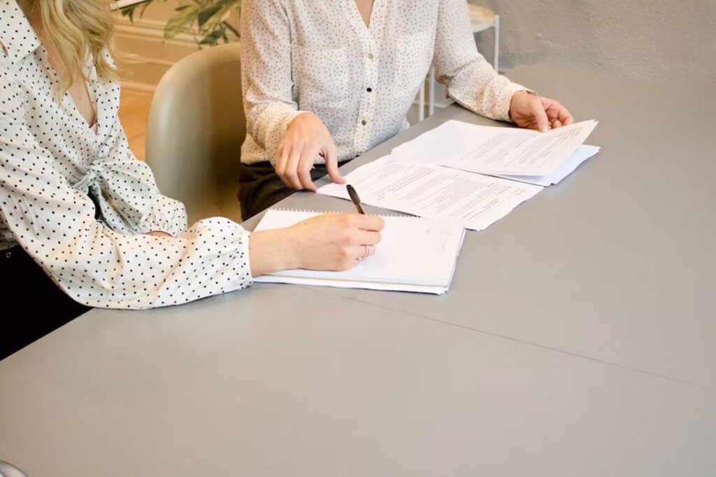 Woman signing a document while another woman is leafing through documents