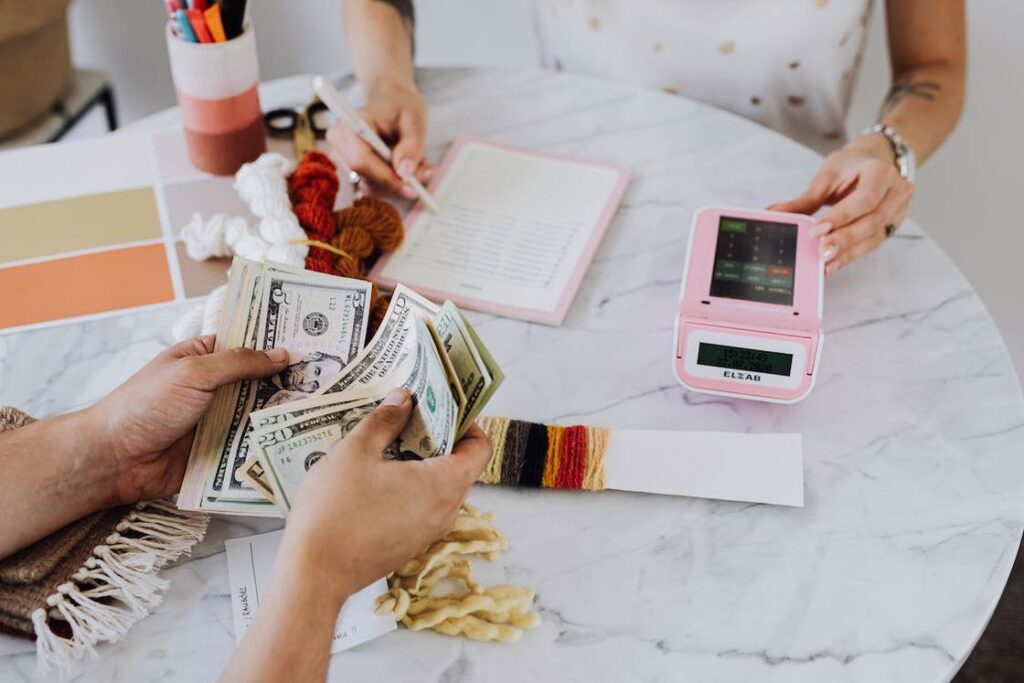 Hand counting dollar bills while another woman is seen writing notes as she uses the calculator