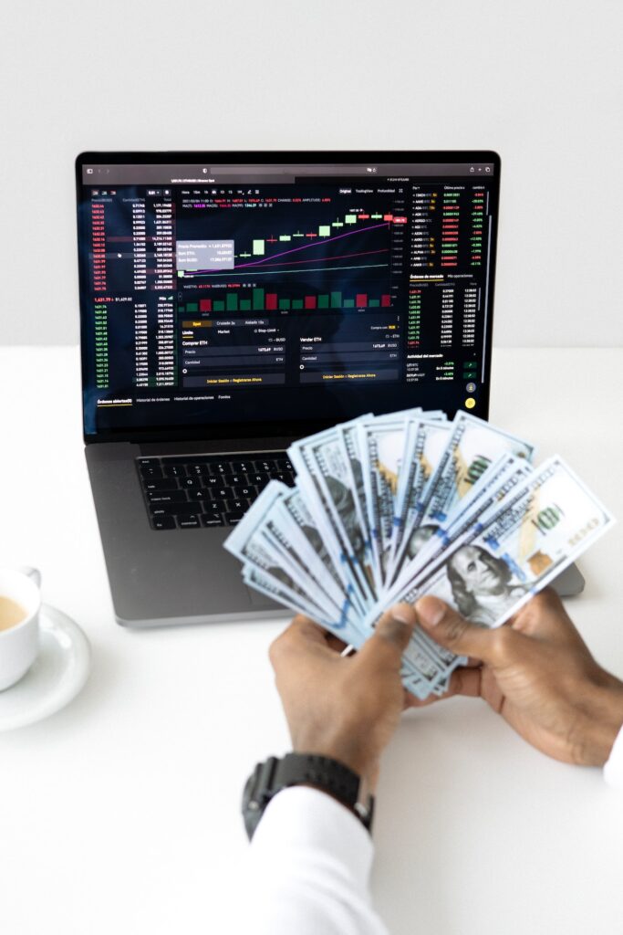 A person holding dollar bills while monitoring the stock market on a silver laptop