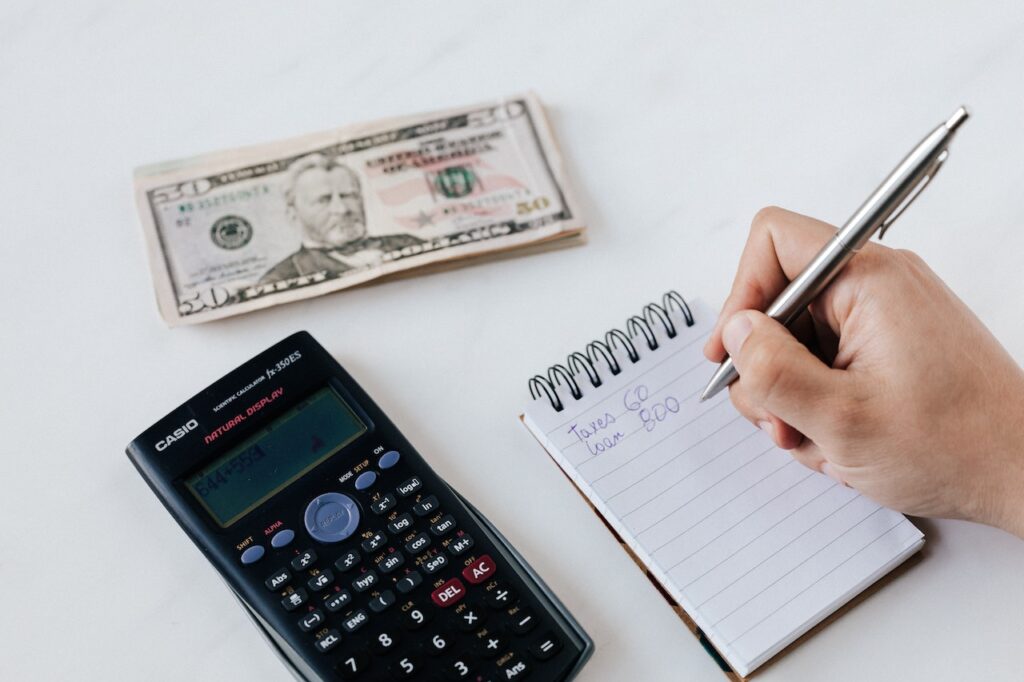 A ballpen used by a person to write on a mini notebook beside a calculator and money