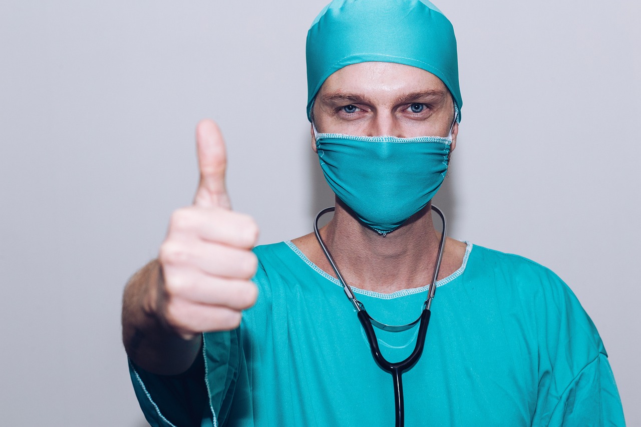 A male surgeon wearing a scrub suit with a stethoscope gives a thumbs up