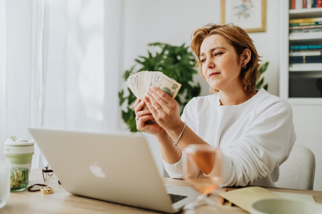 A woman in white long sleeves holding a bunch of cash in front of a silver laptop