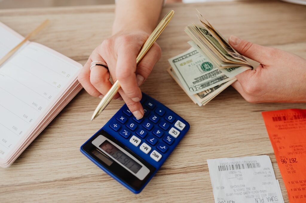 A person holding US dollar bills and a ballpen uses a blue calculator placed on a brown table