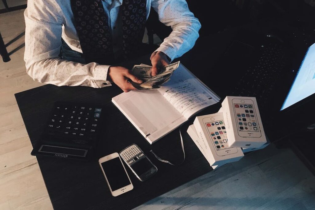 Man counting dollar bills while a calculator, notebook and boxes of iPhones are placed on his table