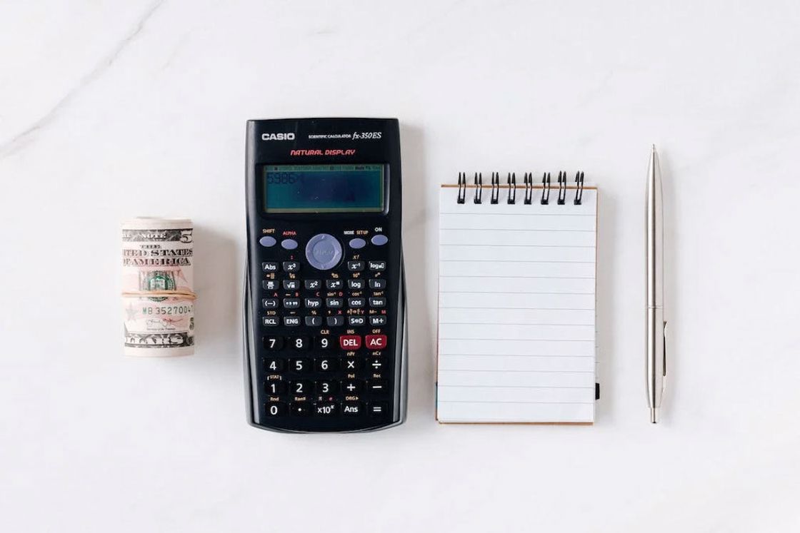 Dollar bills with rubber band, calculator, notepad and pen placed on a white table