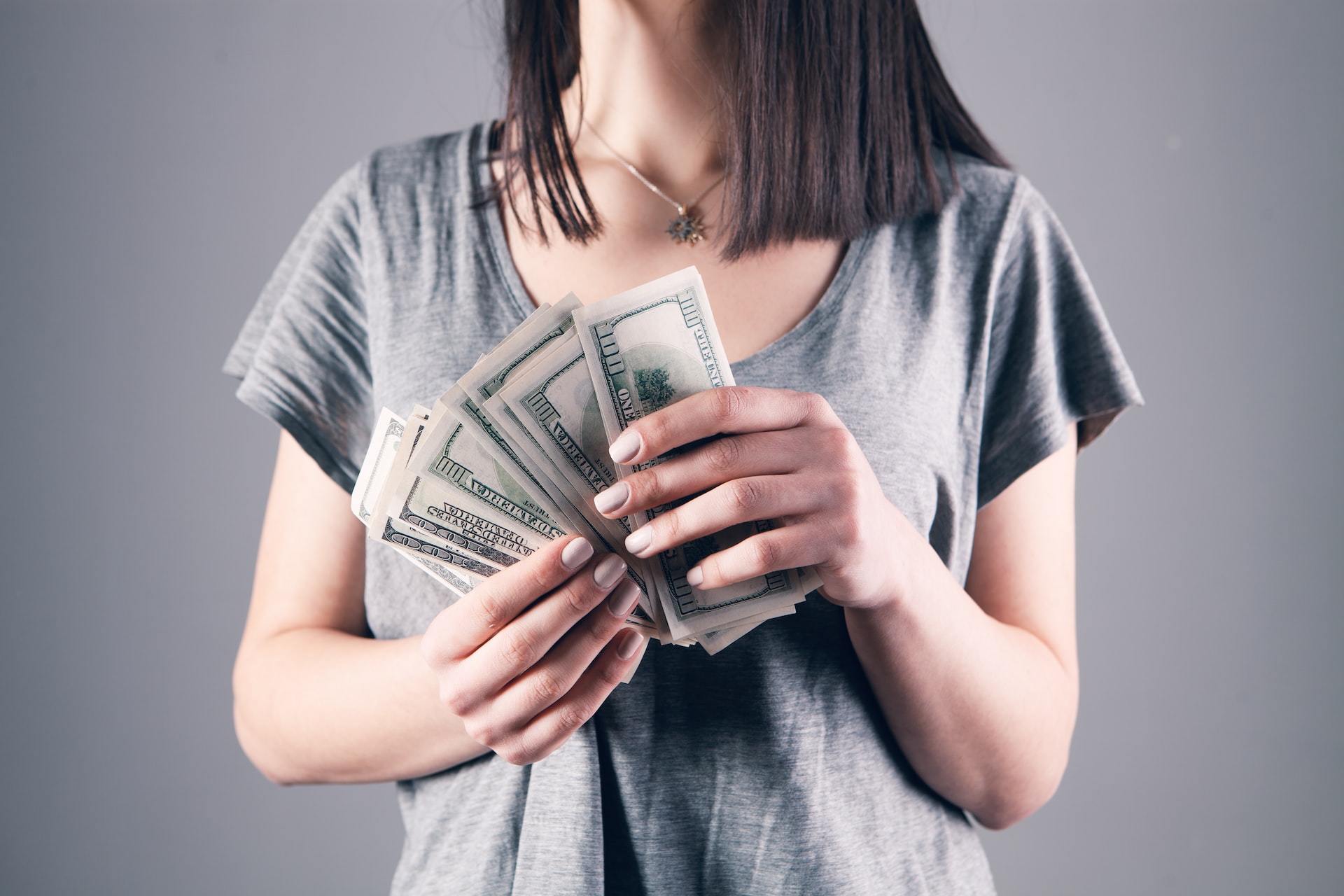 A woman in a gray shirt wearing a necklace holding US dollar bills
