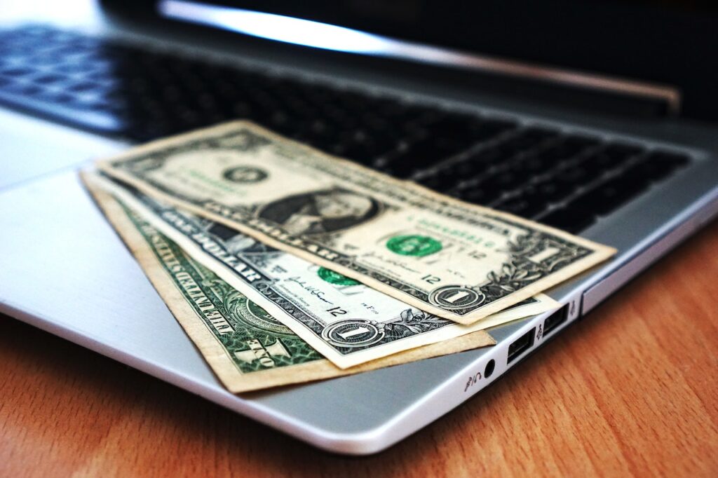 US dollar bills on top of a silver laptop placed on a brown wooden table