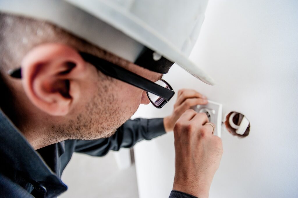 An electrician wearing white hardhat and black glasses is fixing a socket on the white wall