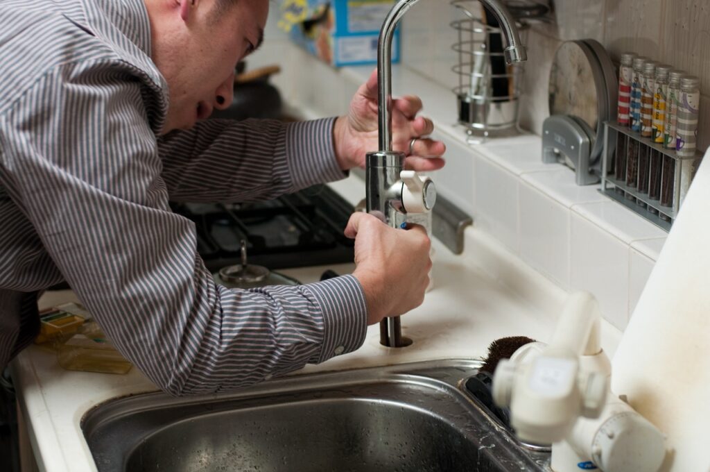 A plumber wearing a striped long-sleeve shirt is fixing the stainless faucet near the kitchen tiles