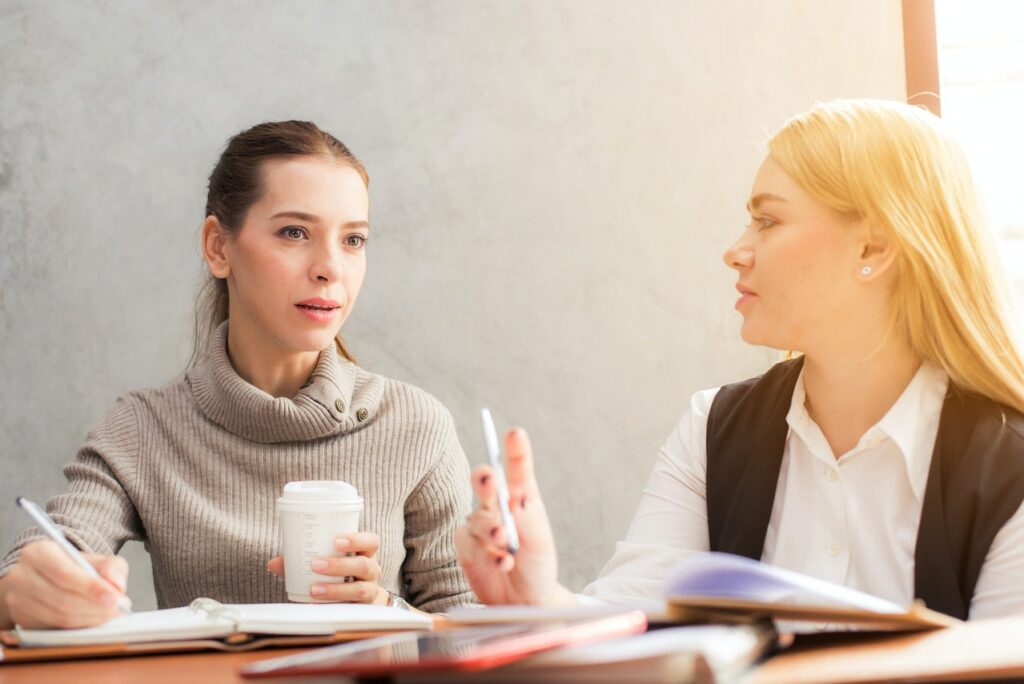 A woman wearing a brown turtleneck is holding a white cup of coffee while discussing with a woman with blonde hair