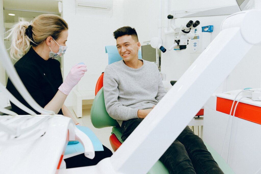 A man wearing a gray sweatshirt and black pants is sitting on a dental chair while smiling at the dental hygienist