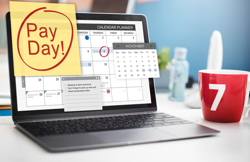 A laptop showing a calendar that says payday beside a red mug
