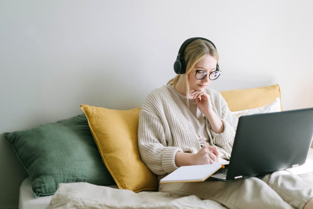 A woman sitting on her bed with her headphones on and laptop open and writing something on a notebook
